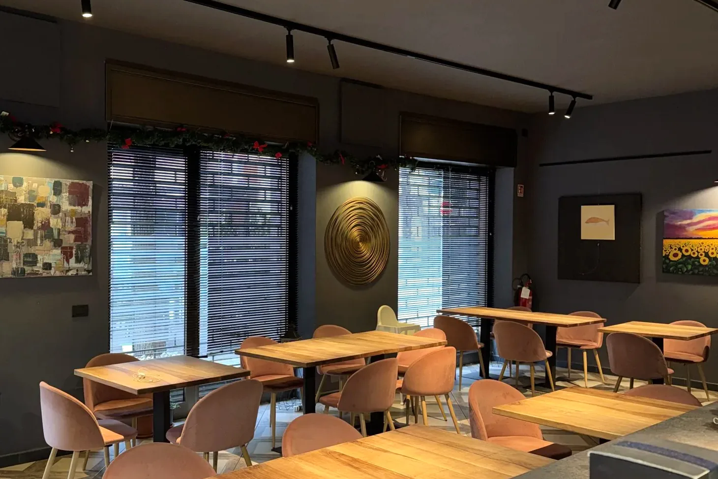 La sala del Nozza: tavoli in legno, sedie color cipria, pareti scure con quadri e luci a binario.